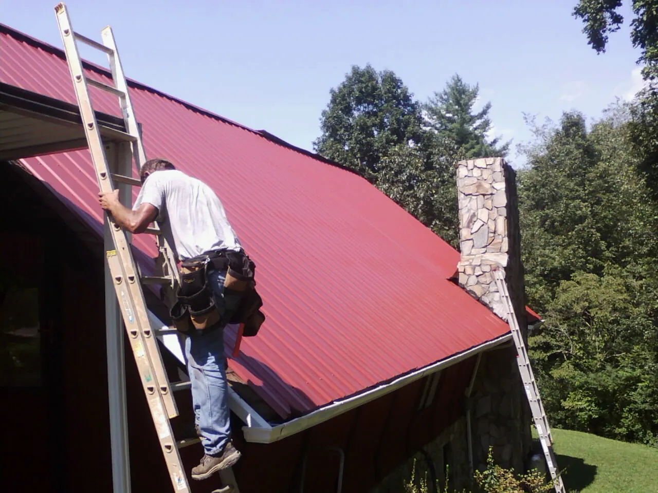 Roofer on a ladder beside a red metal roof and stone chimney — Soffit & Fascia Repair by Roofing Brothers in Leonard